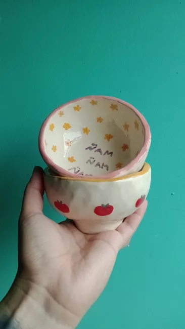 Una mano de tez clara sosteniendo dos bowls de cerámica, uno con tomates dibujados y el otro con flores pequeñas. Fondo turquesa